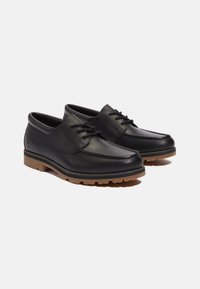 Zapatos de cuero negro con puntera redondeada, que cuentan con cordones y suelas de goma en contraste color tan. Acabado texturizado con detalles de costura.