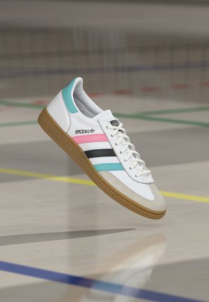 Basket blanche Adidas Spezial avec des bandes roses, noires et bleues, semelle en gomme et bout en daim sur un sol de terrain intérieur.