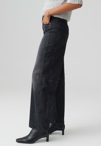 Jeans neri in denim a vita alta e gamba larga con tasche frontali e cuciture sottili. Abbinati a stivali neri con tacco a punta. Texture morbida.