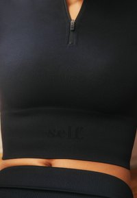 Crni rebrasti sportski crop top s prednjim zatvaračem i suptilnim logotipom na dnu. Čini se da je materijal rastezljiv i prilagođen tijelu.