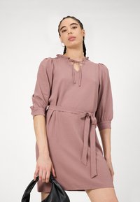 Haljina u cvjetnom uzorku u boji mauve s napućenim rukavima, izrezom u obliku ključa i pojasom vezanim u struku. Model drži crnu torbu.