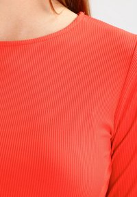 Röd ribbad textil i närbild, med en jämn yta och en båthalsringning. Tyget är elastiskt och formar sig efter kroppens konturer.