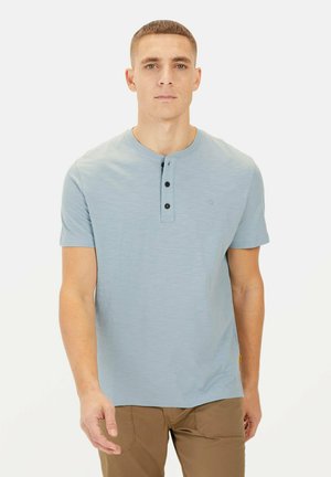Lyseblå kortærmet t-shirt med rund halsudskæring og tre sorte knapper foran, lavet af blødt materiale, parret med brune bukser.