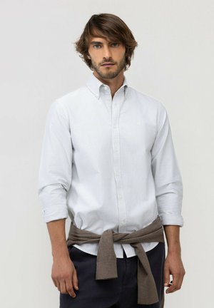 Hombre con cabello castaño medio que lleva una camisa blanca de botones con rayas, pantalones oscuros y un suéter marrón atado alrededor de su cintura contra un fondo liso.