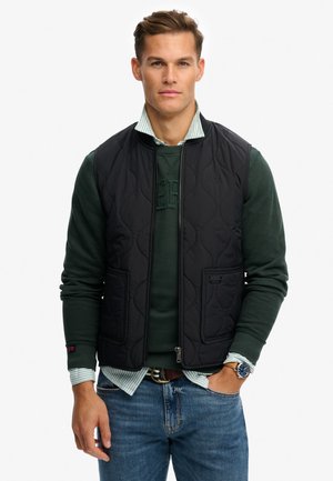 Ung mand iført en sort quiltet vest over en mørkegrøn sweater og en stribet skjorte, blå jeans og et armbåndsur, stående med den ene hånd i lommen.