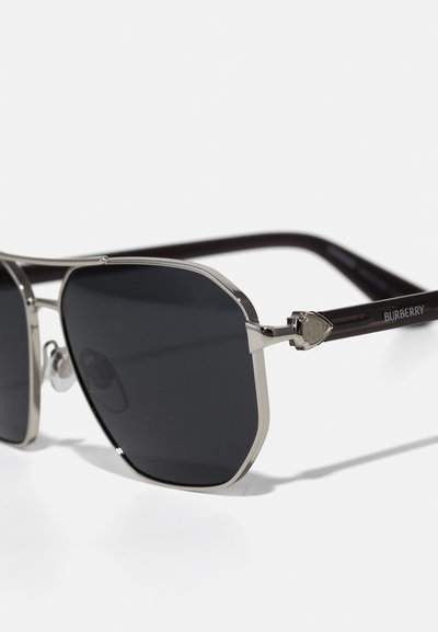 Burberry saulesbrilles ar melniem stikliem, sudraba metāla rāmjiem un tumši brūnām acetāta kājām, kas rotātas ar logotipu un dekoratīvu saspraudes detaļu.