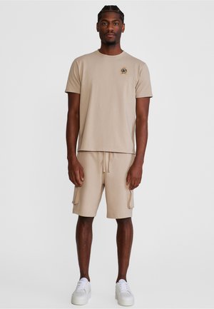 Hombre con camiseta beige con pequeño emblema de escudo, pantalones cortos cargo a juego y zapatillas blancas, de pie frente a un fondo claro y liso.