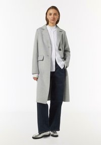 Lichtgrijze wollen jas met een notch-kraag, twee knopen en zijzakken, gelaagd over een wit shirt en donkere, losse broek.