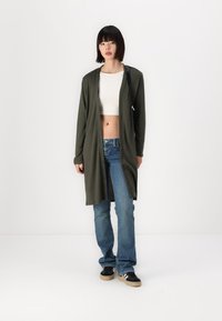 Lange olijfgroene cardigan gemaakt van zachte stof, met een open ontwerp, gecombineerd met een witte crop top en blauwe jeans. Model draagt zwarte sneakers met een beige zool.