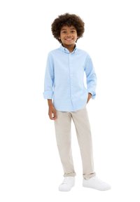 Chemise bleu clair à boutons, manches longues, pantalon beige et baskets blanches ; enfant aux cheveux bouclés souriant, debout avec une main sur la hanche.