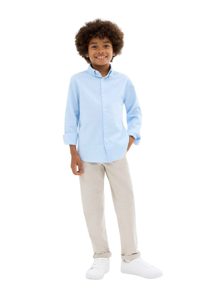 Chemise bleu clair à boutons, manches longues, pantalon beige et baskets blanches ; enfant aux cheveux bouclés souriant, debout avec une main sur la hanche.