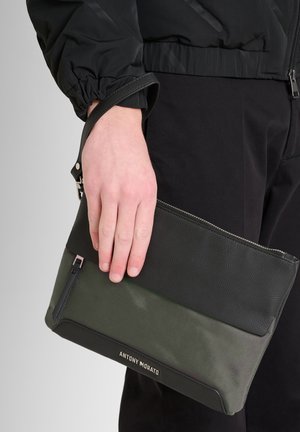 Bolso de mano negro y verde con parte superior de cuero texturizado y sección inferior verde lisa, que cuenta con una correa para la muñeca y cierre de cremallera.