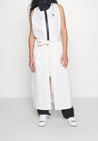 Vit ärmlös klänning med svart dragkedja och mock-halsringning, utrustad med ett knutet midjebälte. Vit textlogotyp på bröstet, designad med en lång, rak skärning.