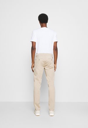 Uomo in piedi, di spalle, che indossa una maglietta bianca a maniche corte, pantaloni beige e sneakers bianche, su uno sfondo chiaro e semplice.