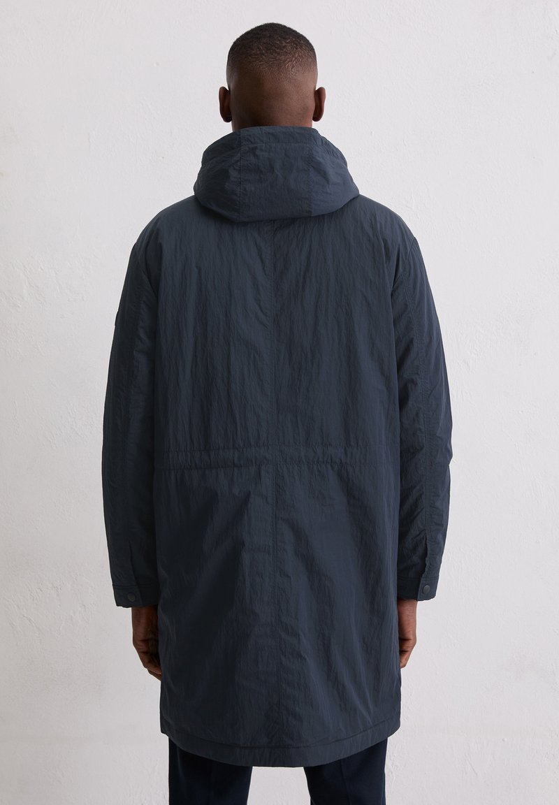 Homme de dos portant un long manteau à capuche bleu marine foncé, debout contre un mur blanc texturé uni.