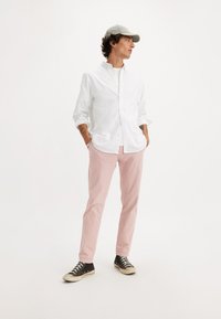 Camicia bianca a maniche lunghe con bottoni, pantaloni rosa chiaro e sneakers in tela nere con suole bianche, abbinati a un cappellino grigio chiaro.