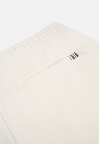 Krämfärgade sweatpants med elastiskt midjeband. Har en enda bakficka med en blå, röd och vit randig detalj. Slät textur.