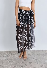 Gonna maxi trasparente nera con motivo di testo scritto a mano bianco, legata in vita, tessuto fluido, lunghezza alla caviglia, texture leggera.