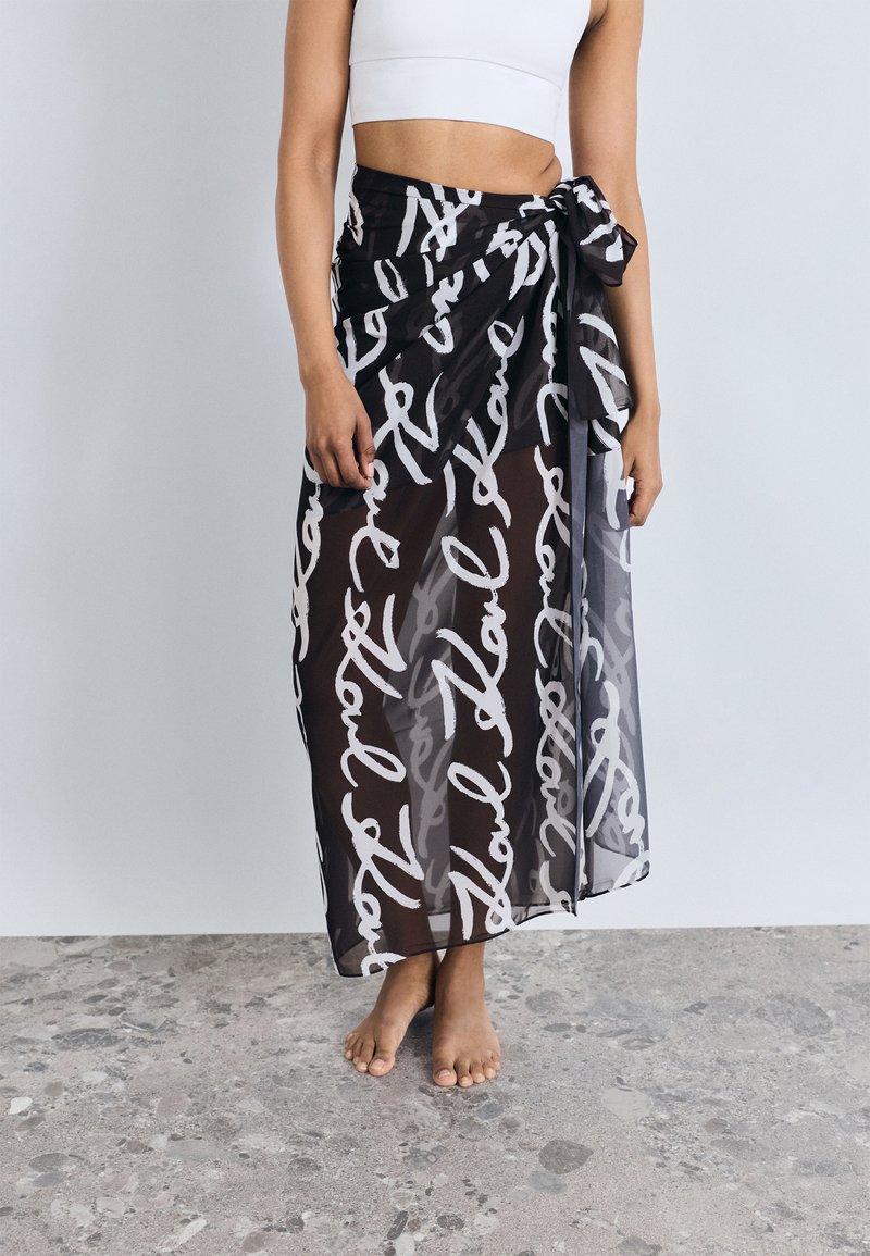 Gonna maxi trasparente nera con motivo di testo scritto a mano bianco, legata in vita, tessuto fluido, lunghezza alla caviglia, texture leggera.