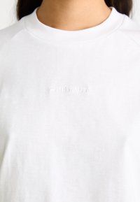 Hvid bomulds t-shirt med rund hals og raglanærmer. Brandlogoet er subtilt præget på fronten for et rent udtryk.
