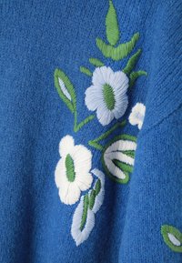 Țesătura tricotată de culoare albastră are broderie florală în alb și albastru deschis, cu frunze verzi. Textura pare moale și texturată.