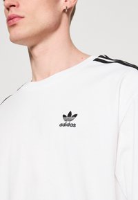 Balta medvilninė marškinėlių su juodu išsiuvinėtu Adidas logotipu krūtinėje ir juodomis juostelėmis ant pečių.