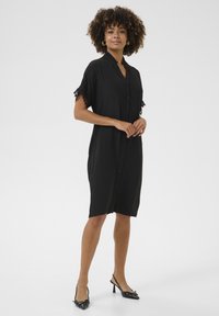 Melna pogu kleita ar augstu apkakli, īsām piedurknēm ar mežģīņu apdari, brīvu piegriezumu un ceļgalu garuma rāvējslēdzēju. Valkāta ar melnām slingback papēžkurpēm.