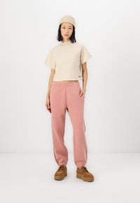 T-shirt beige tagliata con maniche corte, abbinata a pantaloni della tuta rosa chiaro con tasche laterali e polsini elasticizzati. Scarpe platform marroni.