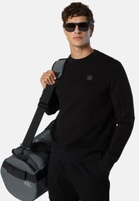 Felpa nera con tessuto strutturato, caratterizzata da un piccolo logo. Borsa da viaggio grigia etichettata "45L" con tracolla. Occhiali da sole indossati.