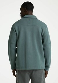 Lichtblauwe pulloverjack met een gestructureerde kraag, textuurstof en contrasterende stikseldetails. Het heeft een ontspannen pasvorm en lange mouwen.