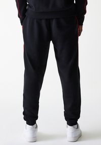 Sorte joggebukser med røde sidestreker, elastisk midje og mansjetter. Tøyet ser mykt og glatt ut; brukt med hvite joggesko.