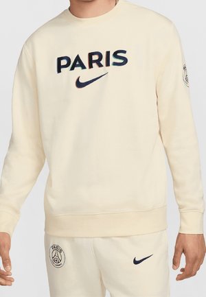 Mężczyzna ubrany w kremową bluzę i spodnie z tekstem "PARIS" oraz logo Nike, a także z emblematami klubu piłkarskiego Paris Saint-Germain na rękawie i spodniach.