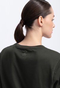 Donkergroene katoenen t-shirt met een ronde halslijn en korte mouwen. Heeft een subtiele schaarafbeelding op de bovenrug. Zachte textuur.