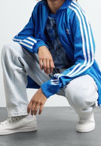 Modrá sportovní bunda s bílými pruhy, na níž je navrstvená modrá košile s vzorem. Světle šedé volné džíny a bílé tenisky dokončují celý outfit.