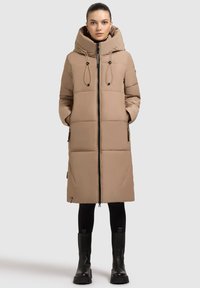 Polstret beige frakke med høj krave, lynlås foran, snore til at stramme, sidelommer og sorte lynlåsdetaljer. Bæret over sorte leggings og støvler.