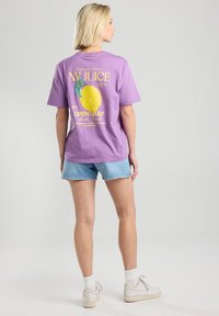 Purple cotton t-shirt featuring a lemon graphic and text on the back. Paired with light denim shorts and white sneakers.