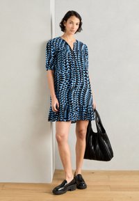 Abito blu e nero a maniche corte con un motivo a fantasie e orlo sfilacciato, abbinato a una borsa tote nera plissettata e scarpe nere chunky.