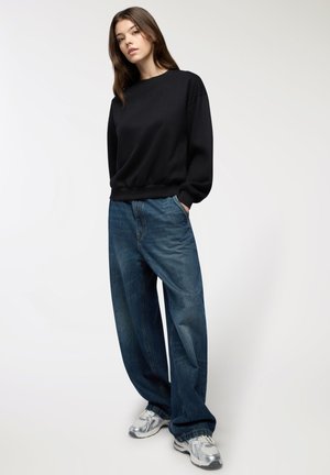 Giovane donna in piedi con le mani nelle tasche, che indossa un felpa nera, jeans blu larghi e scarpe da ginnastica argento su uno sfondo neutro.