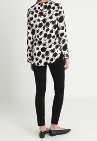 Blouse à manches longues avec un fond blanc et des points abstraits noirs et marron. Associée à un pantalon slim noir et des chaussures plates.