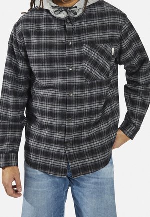 Man wearing black and gray plaid button-up shirt over a gray hoodie paired with light blue jeans, hands relaxed at sides.