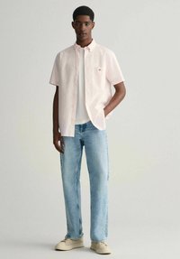 Camicia a maniche corte a quadri rosa chiaro, t-shirt bianca sotto, jeans blu e scarpe beige, esposti su uno sfondo neutro.