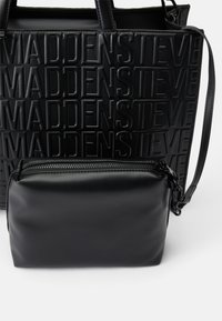Sort håndtaske med præget tekstdesign, der har "MADDEN STEVE." Inkluderer en aftagelig lille clutch med lynlås. Glat tekstur.