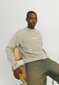 Sudadera gris suave con logo blanco en el pecho, cuello redondo y puños acanalados, combinada con jeans azules. Modelo sentado en una silla de madera.