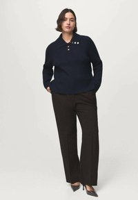 Donna in un maglione blu navy con bottoni dorati, pantaloni larghi neri e décolleté neri con punta, in piedi contro uno sfondo chiaro e uniforme.