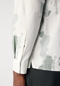 Λευκό πουκάμισο με πράσινα αφηρημένα σχέδια, κουμπωτό μανίκι και αιχμηρή πλάκα μανικιού. Κατασκευασμένο από ένα λείο, ελαφρύ ύφασμα.