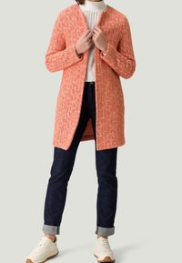Lang cardigan i tekstureret, orange strikmateriale med et åbent frontdesign. Kombineret med mørke jeans og hvide sneakers.