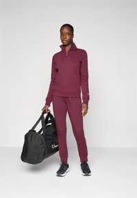 Weinroter Pullover mit halbem Reißverschluss, passende weinrote Jogginghose, schwarze Sportschuhe, hält eine schwarze Reisetasche mit weißem Logo.