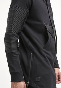 Sudadera negra con textura acanalada en las mangas, detalles de cuero en el bolsillo, cremallera lateral y una etiqueta. Tejido suave, corte alargado y diseño moderno.