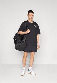 T-shirt oversize noire avec un graphique "ATHLETIC CLUB", associée à un short sombre, des baskets blanches et un sac de sport noir avec une sangle ajustable.
