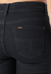 Juodos džinsinės kelnės su užpakaline kišene, pažymėta geltonu "Lois" etikete, kontrastingomis siūlėmis ir prigludusiu dizainu. Tekstūra atrodo lygi.